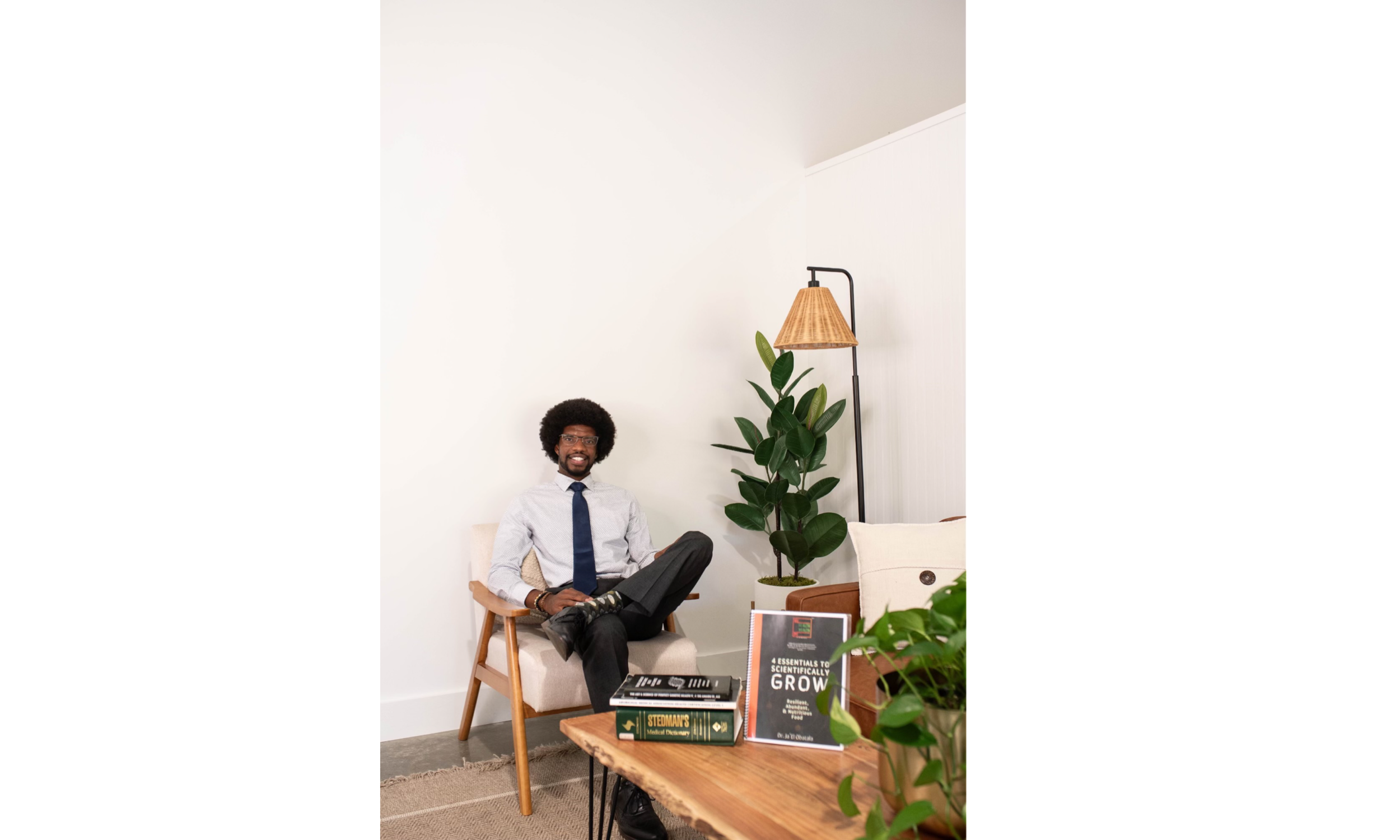Dr. Ja’El Obatala seated beside books and plants